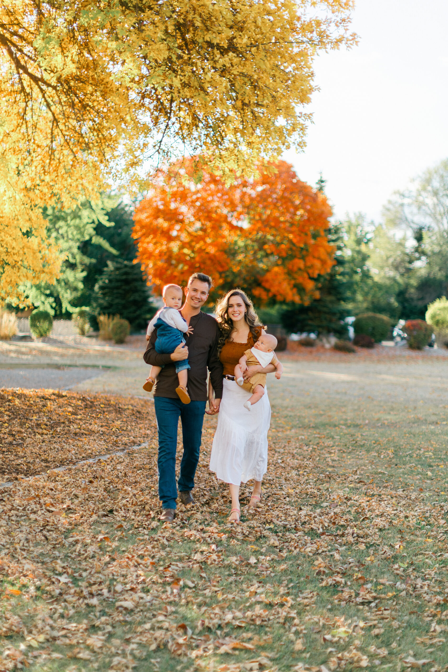 a family of four walking across a field smiling into the camera holding their two little boys with golden light surrounding them and fall leaves in coeur d'alene