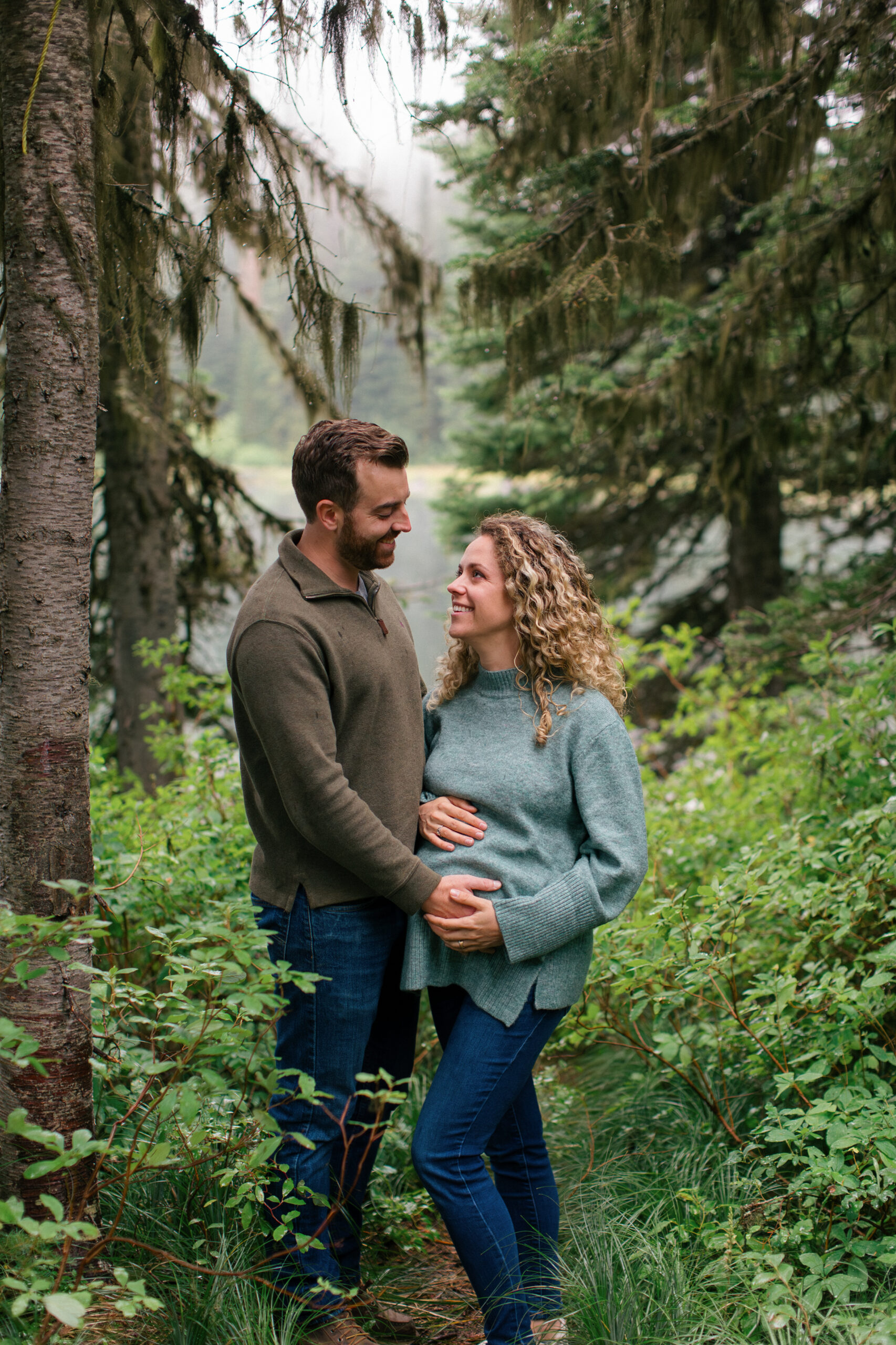An expecting mom and dad rest their hands on her baby bump while smiling at each other, captured during a natural outdoor maternity session in the forest with a misty lake behind them