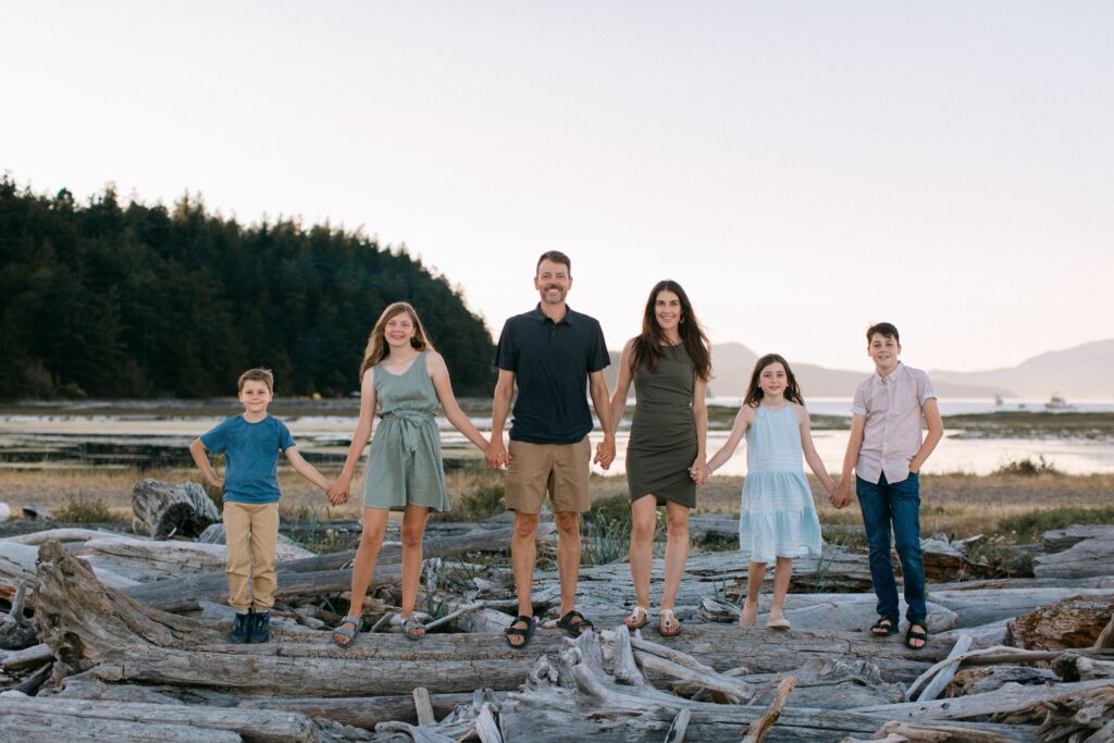 family of six in casual clothes holding hands and walking across a lakeside beach in coeur dalene idaho in the summer 