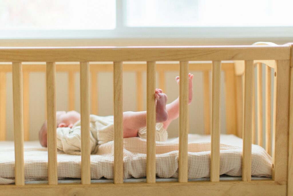 baby boy in crib looking out window with tiny toes peeking through the crib rails
