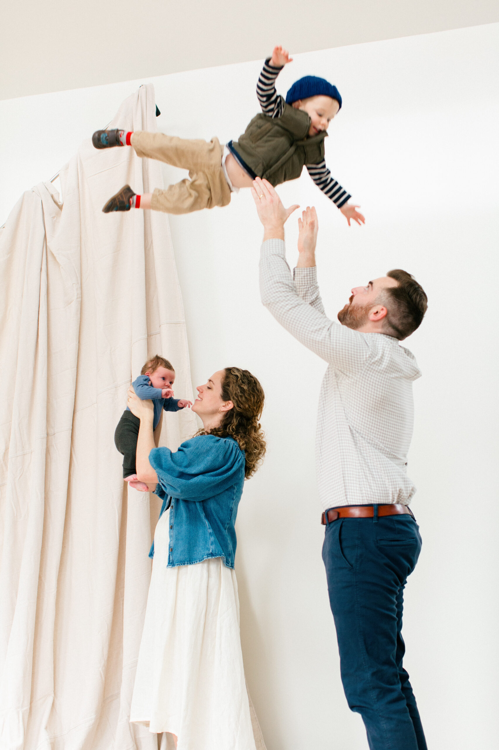 Bright, airy photography studio with tall ceilings, white walls, and natural light streaming in, showing a parents tossing their toddler in the air and playing